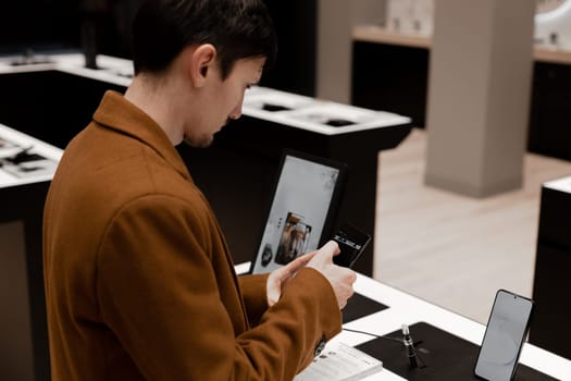 One beautiful young caucasian brunette guy in a brown coat examines a smartphone in a store in a mall, top view close-up. Offline shopping concept.