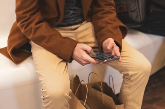 One young caucasian guy in a brown coat and beige trousers flips through a tape in a smartphone while sitting on a plastic white bench in a store in a mall, close-up top view. The concept of using technology, online shopping.