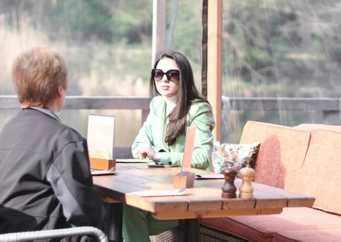 One young beautiful Caucasian psychologist, a brunette girl in a green suit and sunglasses, consults an adult woman sitting on the terrace of a water cafe on a sunny spring day, side view close-up.
