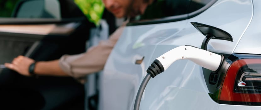 Young man recharge electric car's battery from charging station in outdoor green city park. Rechargeable EV car for sustainable environmental friendly urban travel. Panorama Expedient