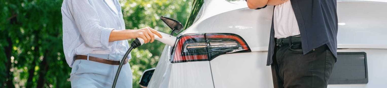 Focused EV car recharging electricity for battery on blurred background of lovey couple ruing their road trip travel by eco friendly electric car in national park and greenery forest on holiday. Exalt