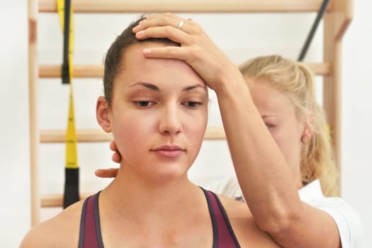 Young physiotherapist exercising with her female patient, fixing head and neck using hand, during exercise