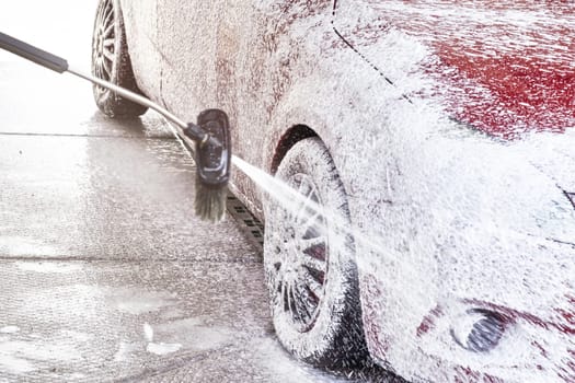 Red car washed in self serve carwash, detail on white soap foam spraying from high pressure hose onto wheel