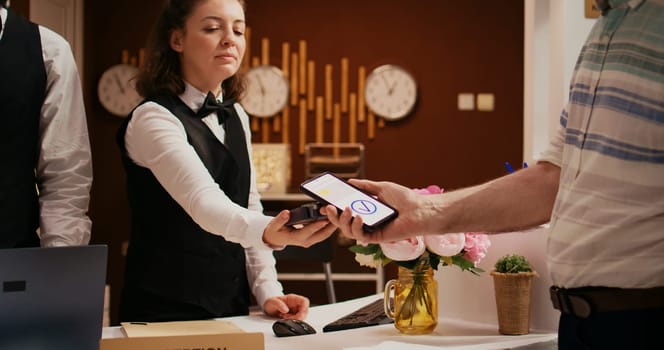 Retired guest using mobile payment with nfc technology at pos terminal, paying for hotel room at reception front desk. Old man registering at holiday retreat, making electronic transaction.