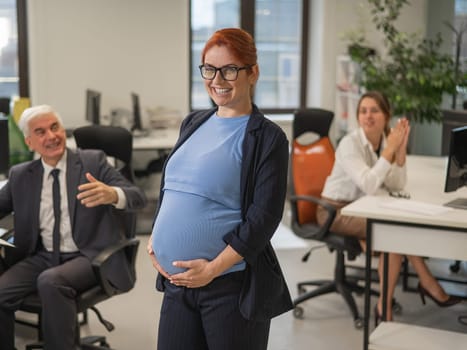 A happy pregnant woman stands in the middle of the office next to a Caucasian woman and an elderly man working at computers