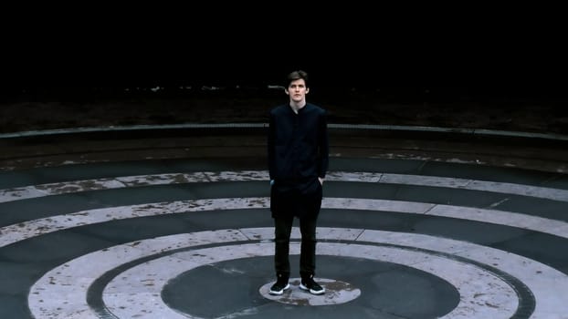 Tall man in black clothes standing in the meddle of amphitheater. Stock clip. Asphalt colored in black circles