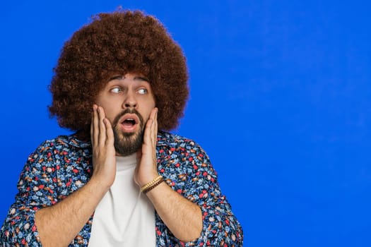 Oh my God, Wow. Amazed winner man with Afro hairstyle wig raising hands in surprise looking at camera with big eyes shocked by victory celebrating success good news. Guy on blue background. Copy-space