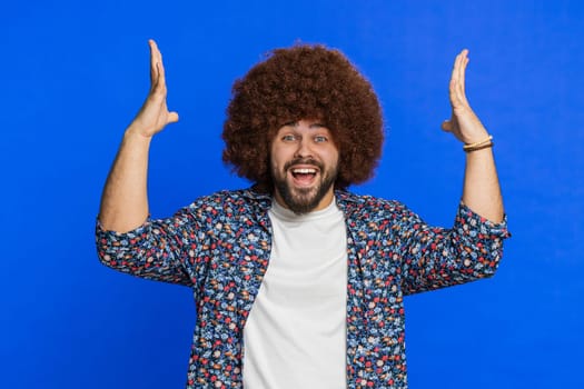 Shocked amazed surprised man with Afro hairstyle wig touching his head showing hands brain explosion wow gesture, professional burnout, celebrating success victory win. Guy isolated on blue background