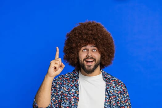 Happy man with Afro hairstyle wig showing pointing up overhead empty place, advertising area for commercial text copy-space for goods promotion advertisement logo text. Guy isolated on blue background