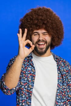 Okay. Happy man with Afro hairstyle wig looking approvingly at camera showing ok gesture, like sign positive feedback, approve something good, celebrate victory. Guy on blue background. Vertical
