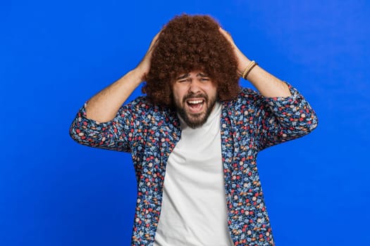 Irritated frustrated man with Afro hairstyle wig screams from stress tension problems feels horror hopelessness fear surprise shock expresses rage nervous, quarrel. Guy isolated on blue background
