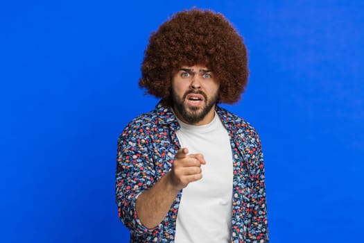 Quarrel. Displeased aggressive unhappy man with Afro hairstyle wig gesturing hands with irritation displeasure blaming scolding for failure asking why this happened. Guy isolated on blue background