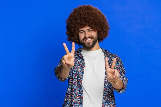 Happy Caucasian man showing victory sign, hoping for success and win, doing peace gesture, smiling with kind optimistic expression, celebrate. Stylish young guy isolated on studio blue background