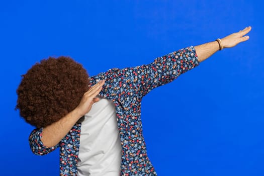 Trendy cheerful positive man with Afro hairstyle wig having fun dancing and moving to rhythm, dabbing raising hands making dub dance winner celebrating victory gesture. Guy isolated on blue background