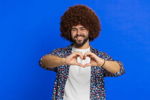 Man in love. Smiling attractive Caucasian man makes heart gesture demonstrates love sign expresses good positive feelings and sympathy. Stylish young lovely boy isolated on studio blue background