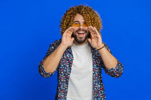 Happy excited Caucasian man yell, shout to camera, promotion advertisement of holidays sale, invitation, greetings, welcome. Excited smiling stylish young guy scream loud isolated on blue background