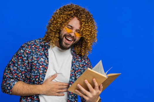 Happy relaxed smiling man with curly hairstyle wig reading funny interesting fairytale story book, leisure hobby, knowledge wisdom, education, learning, study, wow. Guy isolated on blue background