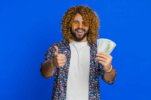 Rich excited happy man with curly hairstyle wig waving money dollar cash banknotes bills like a fan, success business career, lottery winner, big income wealth. Guy isolated on blue background indoors