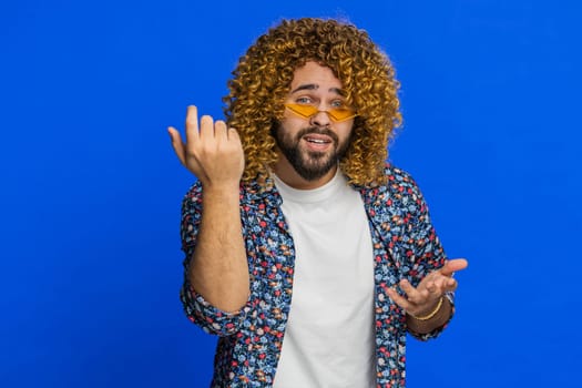 Knock-knock who is there. Confused amazed man with curly hairstyle wig knocking door gesture asking who is at home, feeling embarrassed, no idea, being clueless and uncertain. Guy on blue background