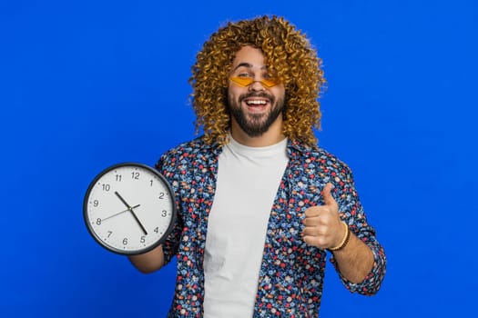 It is your time, hurry up. Caucasian man showing time on wall office clock, ok, thumb up, approve, pointing finger at camera, advertisement, deadline. Stylish young guy isolated on blue background