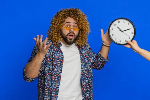 Man with curly hairstyle wig with anxiety checking time on clock running late to work being in delay deadline panic. Guy looking at hour, minutes, worrying to be punctual isolated on blue background