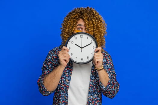 Serious strict man with curly hairstyle wig holding office clock hiding behind, checking time on watch, running late to work, being in delay, deadline, obscuring face. Guy isolated on blue background