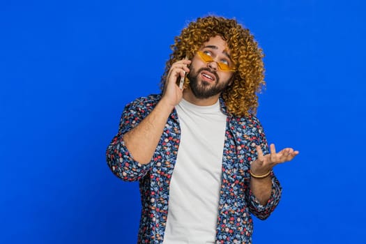 Disinterested man with curly hairstyle wig having annoyed boring talk on smartphone. Guy tired sleepy guy talking on mobile phone with friend, making online conversation on blue background. Copy-space