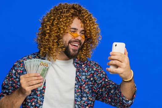 Happy rich man with curly hairstyle wig looking smartphone display sincerely rejoicing win receiving money dollar cash banknotes celebrating success victory play lottery game. Guy on blue background