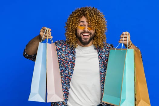Happy Caucasian man showing shopping bags, advertising discounts, smiling looking amazed with low prices, shopping on Black Friday holidays. Stylish young guy isolated on blue background. Copy-space