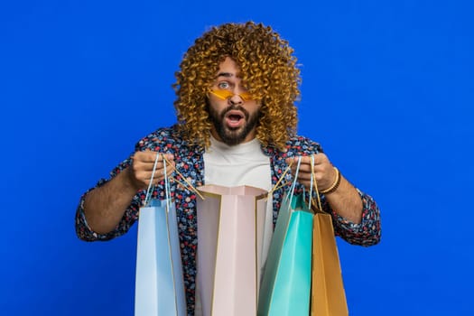 Excited Caucasian man showing shopping bags, advertising discounts, smiling looking amazed with low prices, shopping on Black Friday holidays. Stylish young guy isolated on blue background. Copy-space
