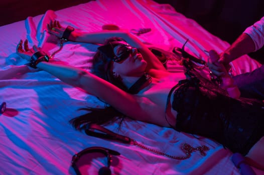A woman in a leather mask and handcuffs lies on a bed in a blue-red light. Man holding dildo