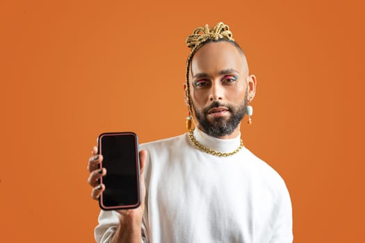 Young smiling african-american gay man wear sweatshirt hold in one hand mobile phone show smartphone screen to camera isolated on orange background studio portrait. Lgbtq pride concept