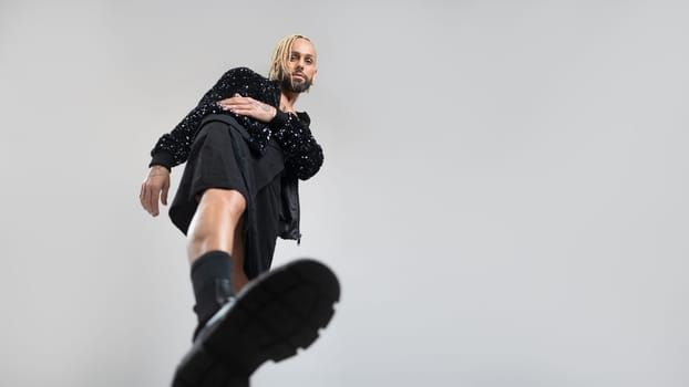 Full length gay man wearing black skirt and big black boots doing step on camera. Afro-american homosexual posing in photo studio on white background. Bearded gay with make up