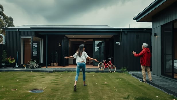 A woman and a child are playing in the backyard of a house, surrounded by green grass and plants. The facade of the building, with a roof and a lawn, overlooks the leisure event, under the cloudy sky