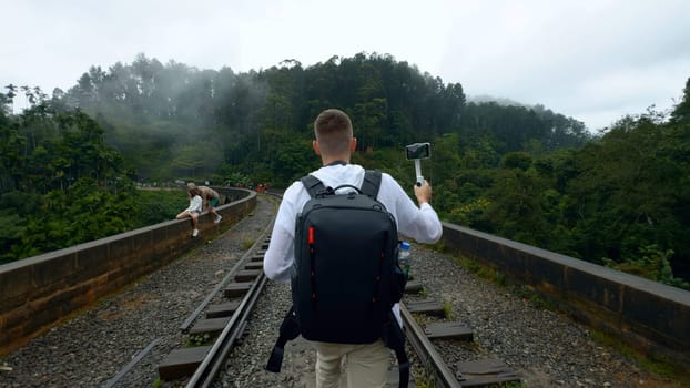 Caucasian traveler man recording video of green mountain landscape. Action. Man walking on railroad with backpack
