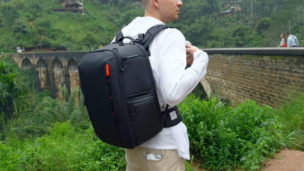 Man in jungle near bridge. Action. Man with backpack looks back at hiking trail in jungle. Handsome man on top of ancient stone bridge in jungle.