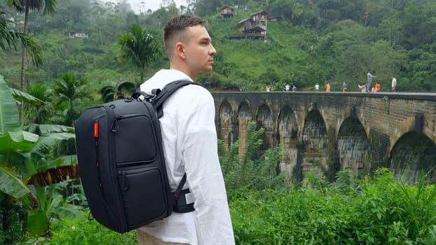 Man in jungle near bridge. Action. Man with backpack looks back at hiking trail in jungle. Handsome man on top of ancient stone bridge in jungle.