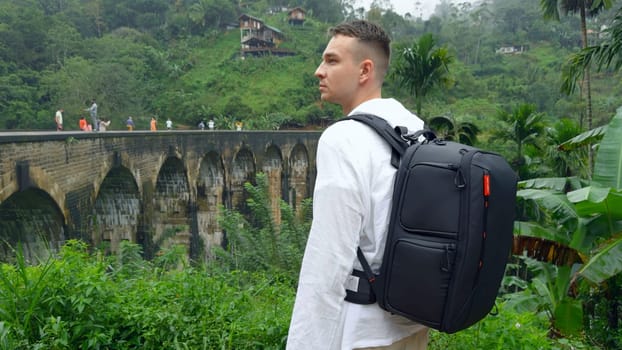 Man in jungle near bridge. Action. Man with backpack looks back at hiking trail in jungle. Handsome man on top of ancient stone bridge in jungle.