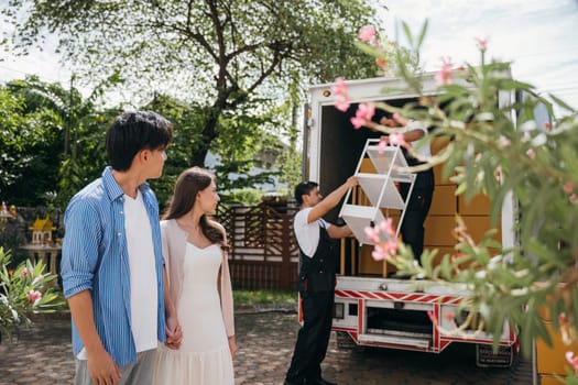 A smiling newlywed couple's portrait while professional movers load furniture onto a truck for relocation. Quality service and teamwork for satisfied customers. Moving Day Concept