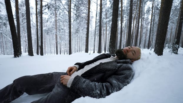 Man lies in snow in winter forest. Media. Tired man lies in snowdrift in winter forest. Depressive Stylish man lies in snow in winter forest.