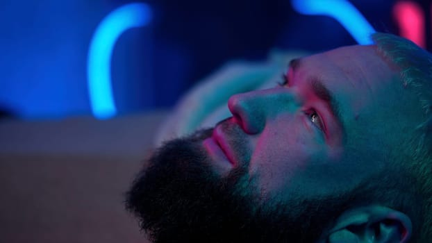 Close up side view of a bearded man looking up and thinking of something. Media. Young man with neon light on the background