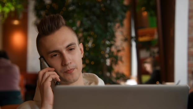 Young man is working on laptop in cafe and talking on phone. Stock footage. Young freelancer is working on laptop and answering calls. Remote work with laptop and cafe calls.