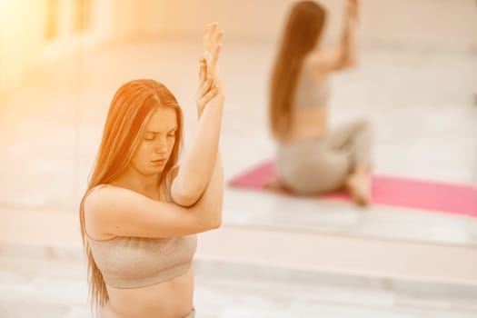 Young woman doing yoga in the gym. A girl with long hair and in a beige tracksuit stands in a cow pose on a pink carpet. A woman performs Gomukhasana