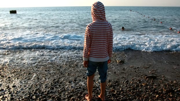 Happy child enjoys the sea on a hot summer day. Creative. Strong waves and boy standing barefoot on stony shore