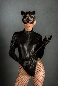 Adult sex games. Beautiful dominant brunette vamp mistress girl in latex body, gloves and bdsm black leather fetish cat mask posing with riding crop. - image