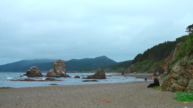 People relax on rocky coast with mountains on cloudy day. Clip. Beautiful coast with rocks and rocks sticking out on shore. People relax on beautiful coast with rocks and mountains.