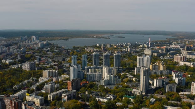 City with high-rise buildings on background of lake and green forest. Stock footage. Modern buildings in beautiful green city with lake and forest horizon. Top view of panorama of modern city in forest area.