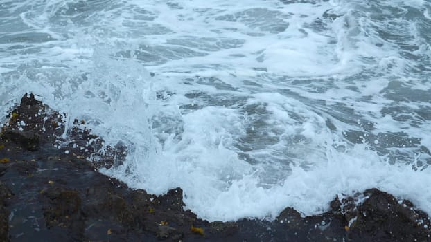 Close-up of surge of waves on rocky shore. Clip. Beautiful waves with splash roll on stones of seashore. Beautiful splashes of waves crashing on rocks on cloudy day.