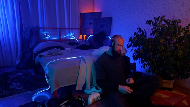 Young man in hipster room with neon lighting listening to music. Media. Man enjoying music with headphones on his head