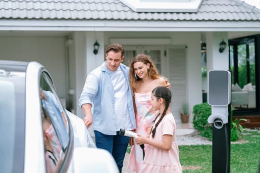 Happy little young girl learn about eco-friendly and energy sustainability as she help her family recharge electric vehicle from home EV charging station. EV car and modern family concept. Synchronos